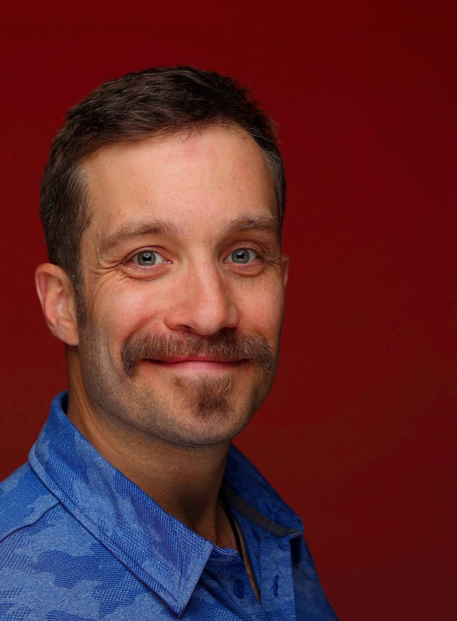 Smiling man with mustache in blue shirt against deep red background Headshot