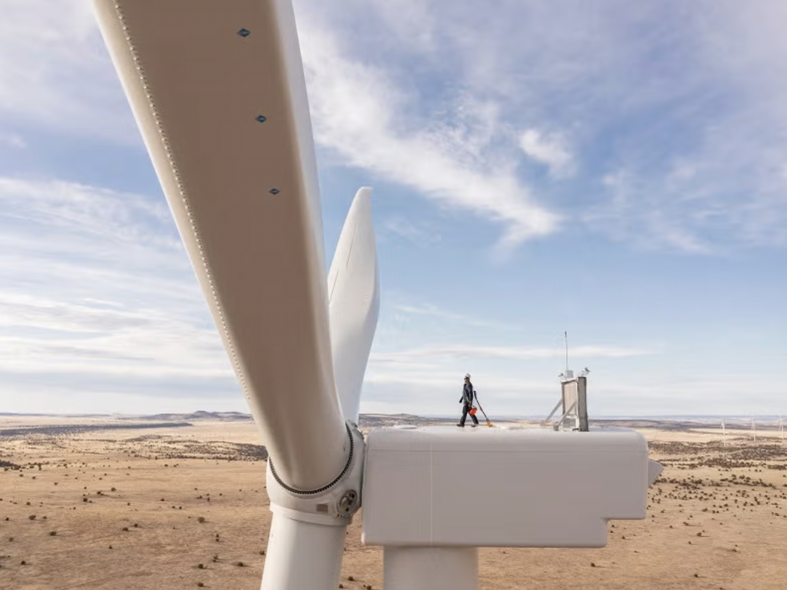 Close-up of white wind turbine blade with technician and control tower visible in arid landscape