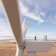 Close-up of white wind turbine blade with technician and control tower visible in arid landscape