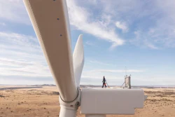 Close-up of white wind turbine blade with technician and control tower visible in arid landscape