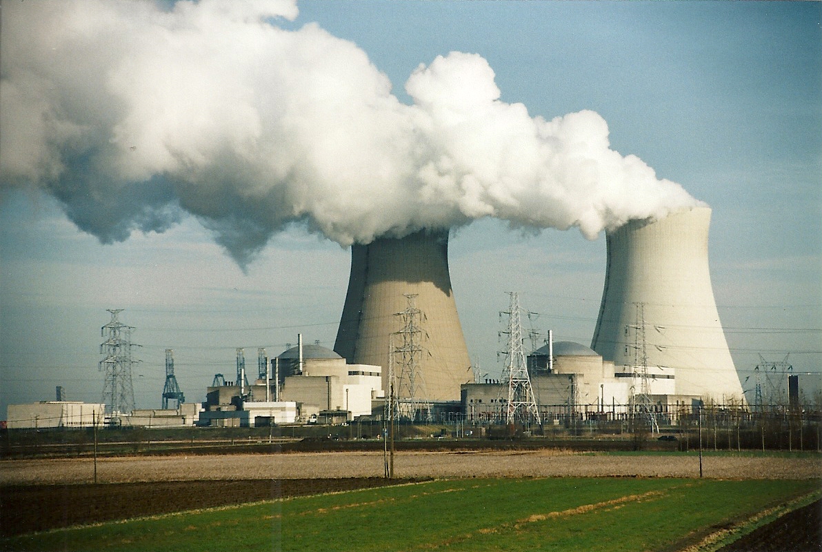 Nuclear power plant with two cooling towers emitting steam, surrounded by electrical transmission towers and industrial facilities against a blue sky.