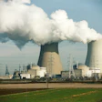 Nuclear power plant with two cooling towers emitting steam, surrounded by electrical transmission towers and industrial facilities against a blue sky.