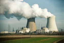 Nuclear power plant with two cooling towers emitting steam, surrounded by electrical transmission towers and industrial facilities against a blue sky.