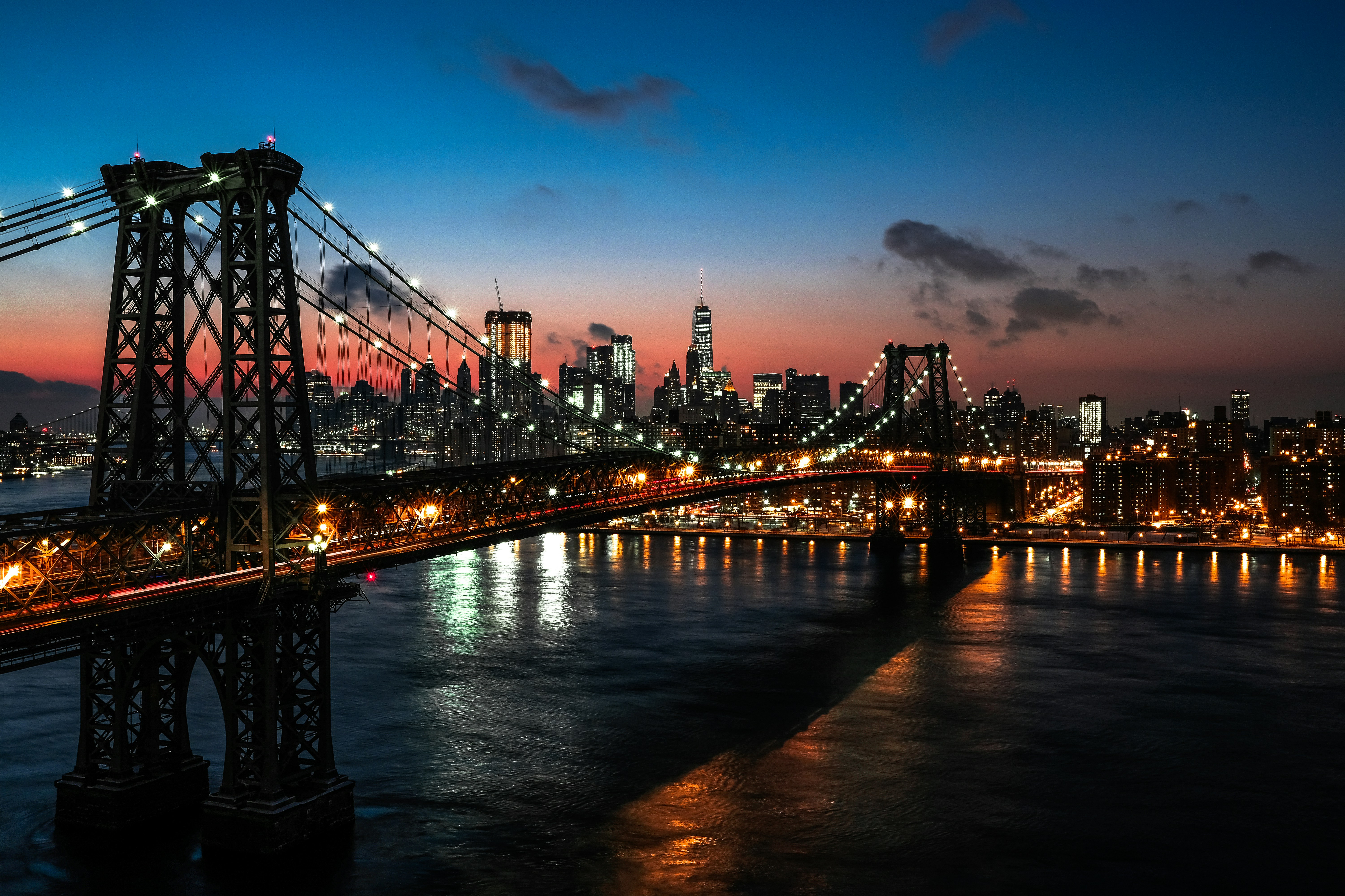 Williamsburg Bridge spanning the East River at dusk with Manhattan skyline illuminated against a gradient sunset sky