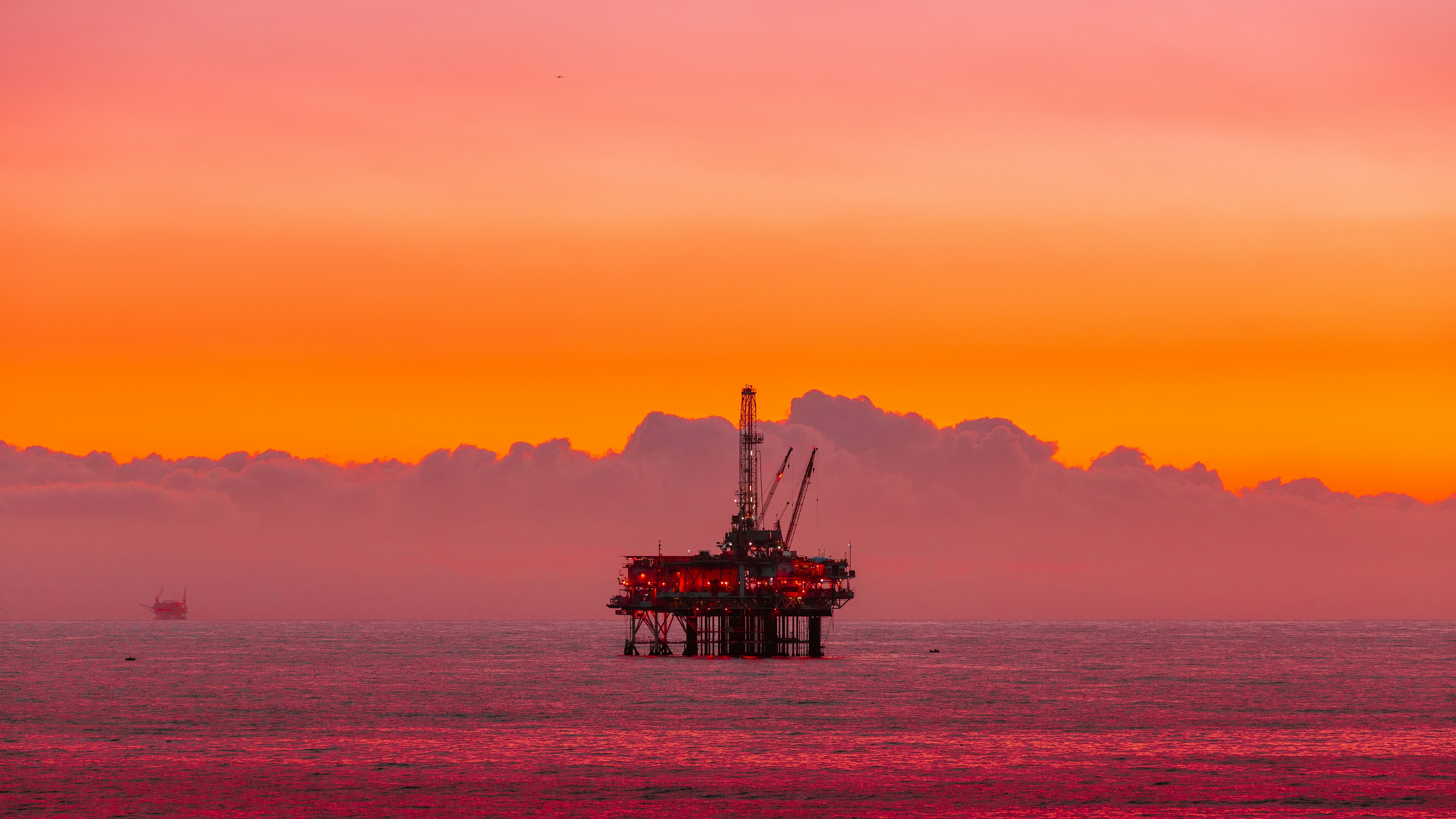 Offshore oil drilling platform silhouetted against vibrant orange and pink sunset sky over calm ocean waters