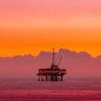 Offshore oil drilling platform silhouetted against vibrant orange and pink sunset sky over calm ocean waters