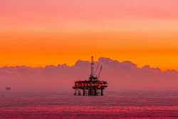 Offshore oil drilling platform silhouetted against vibrant orange and pink sunset sky over calm ocean waters