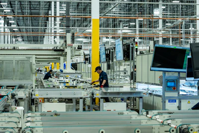 Industrial factory floor with workers assembling components at production stations with yellow structural supports and digital displays