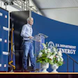 Speaker at podium during U.S. Department of Energy event with blue branded backdrop and floral arrangement