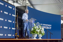 Speaker at podium during U.S. Department of Energy event with blue branded backdrop and floral arrangement