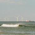 Offshore wind turbines line the horizon beyond breaking waves on a sandy beach, with a cargo ship and seabird visible in the distance