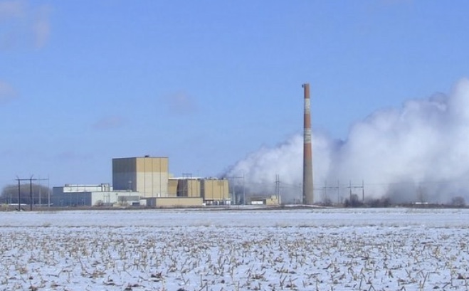 Industrial power plant with tall smokestack emitting steam, surrounded by snowy field under blue sky