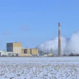 Industrial power plant with tall smokestack emitting steam, surrounded by snowy field under blue sky