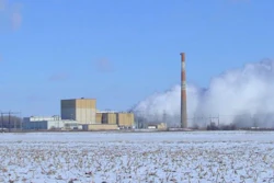 Industrial power plant with tall smokestack emitting steam, surrounded by snowy field under blue sky