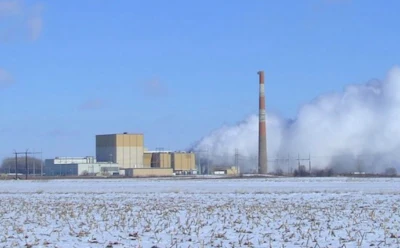 Industrial power plant with tall smokestack emitting steam, surrounded by snowy field under blue sky