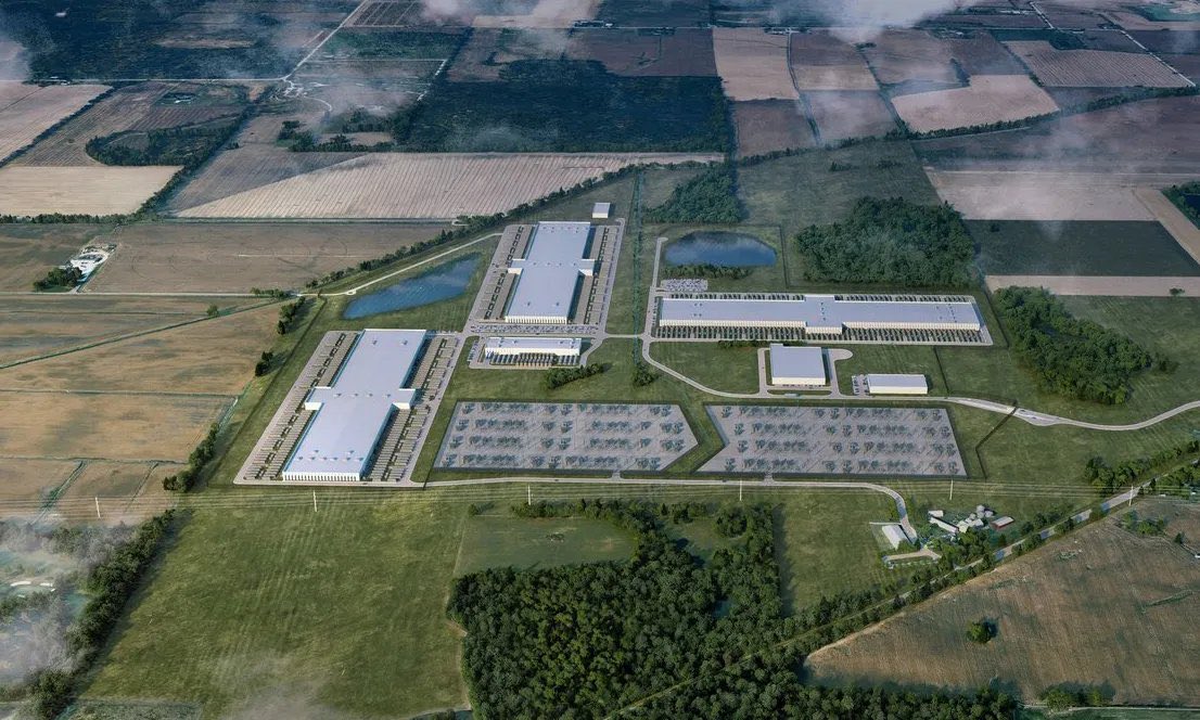 Aerial view of industrial manufacturing facility with warehouses, solar panels, and parking areas surrounded by agricultural fields