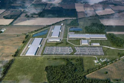 Aerial view of industrial manufacturing facility with warehouses, solar panels, and parking areas surrounded by agricultural fields