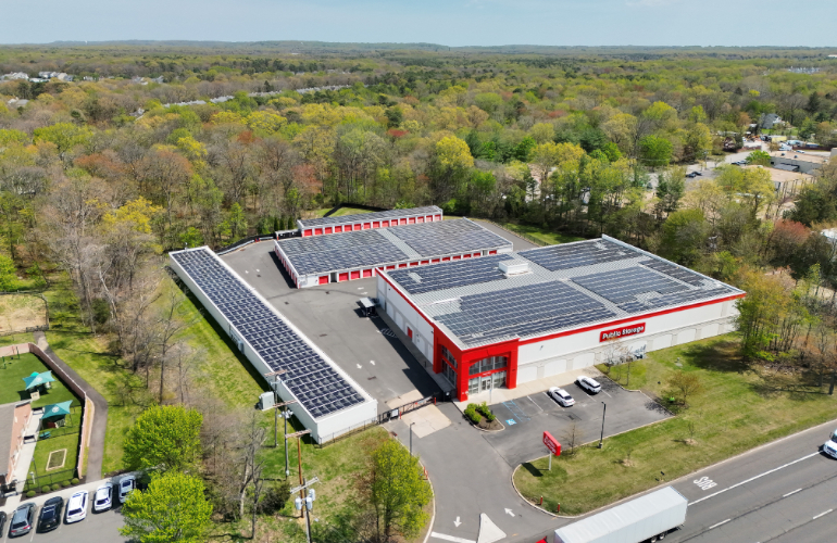 Aerial view of red and white industrial facility with extensive solar panel arrays surrounded by forested landscape