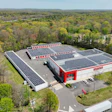 Aerial view of red and white industrial facility with extensive solar panel arrays surrounded by forested landscape