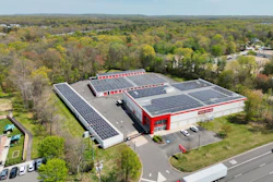 Aerial view of red and white industrial facility with extensive solar panel arrays surrounded by forested landscape