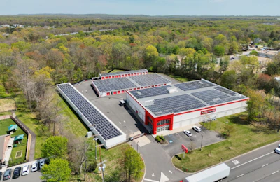 Aerial view of red and white industrial facility with extensive solar panel arrays surrounded by forested landscape