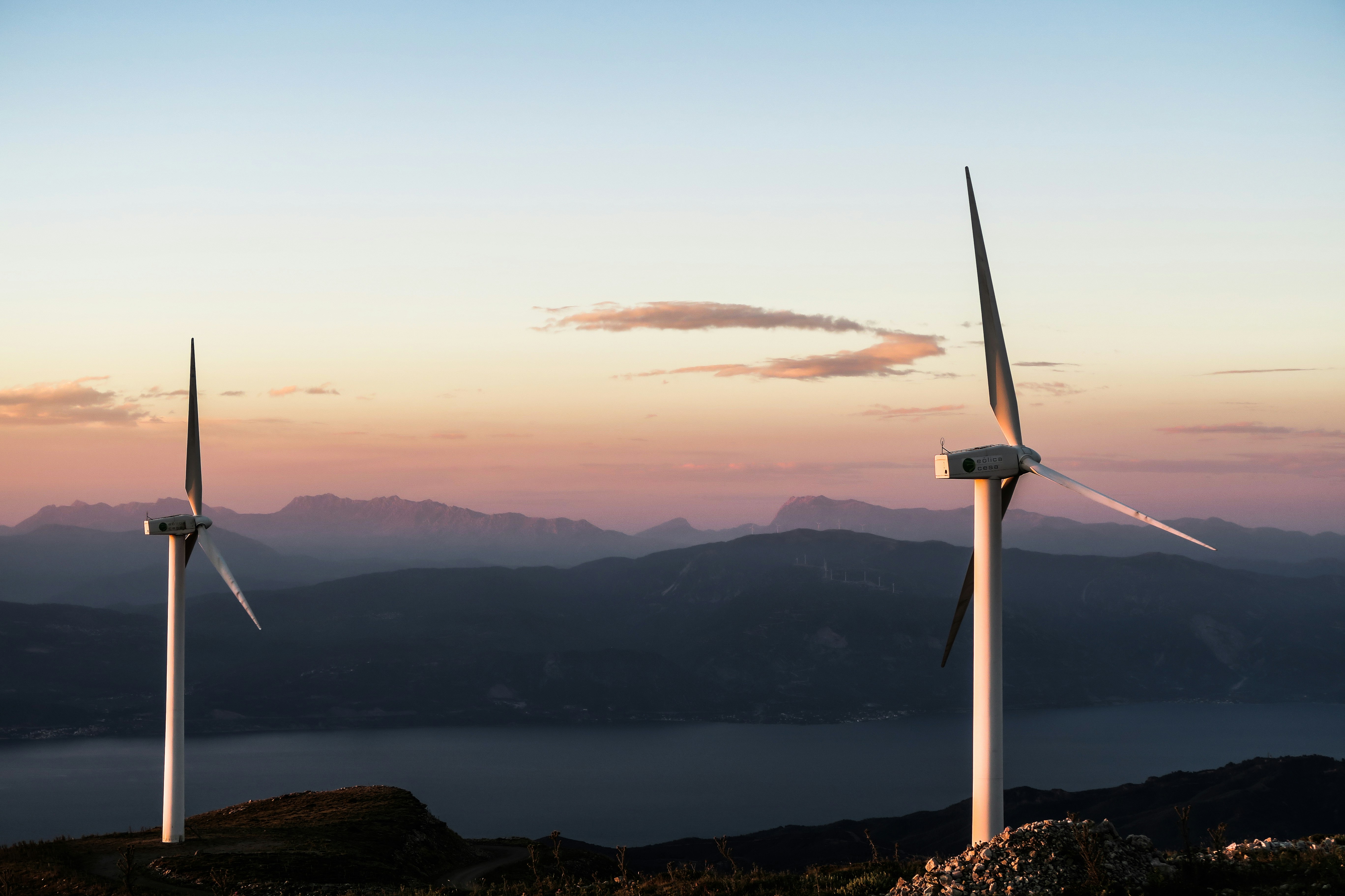 Two wind turbines silhouetted against a twilight sky with mountains and a lake in the background