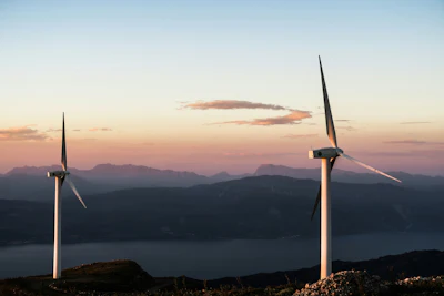 Two wind turbines silhouetted against a twilight sky with mountains and a lake in the background